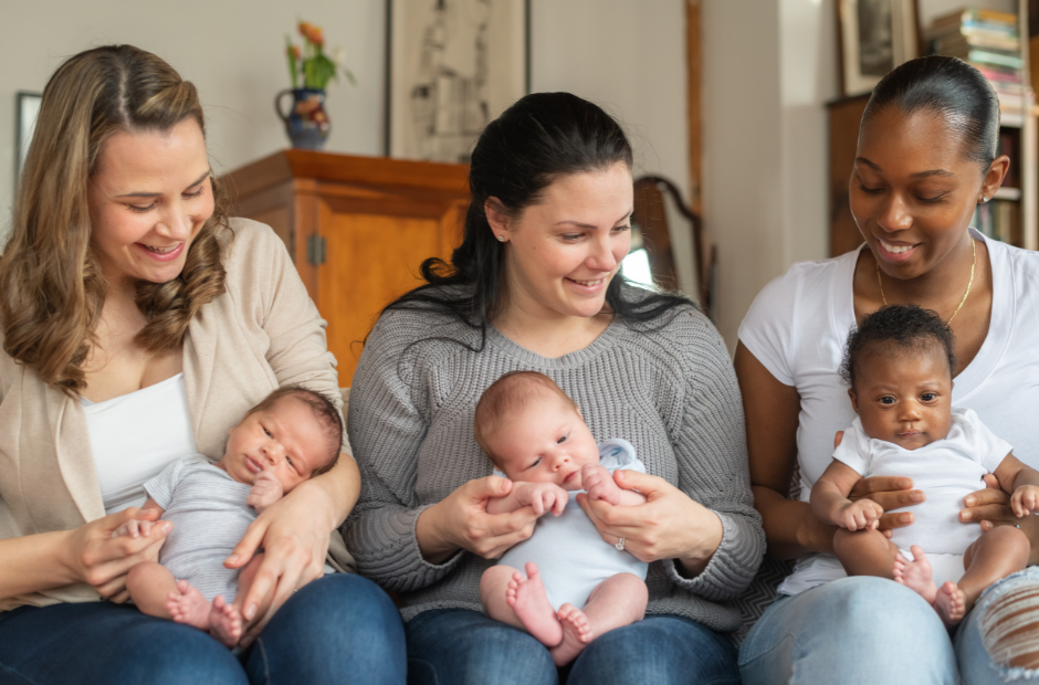 Three women sit in a row, holding new born babies on their laps. They are smiling.